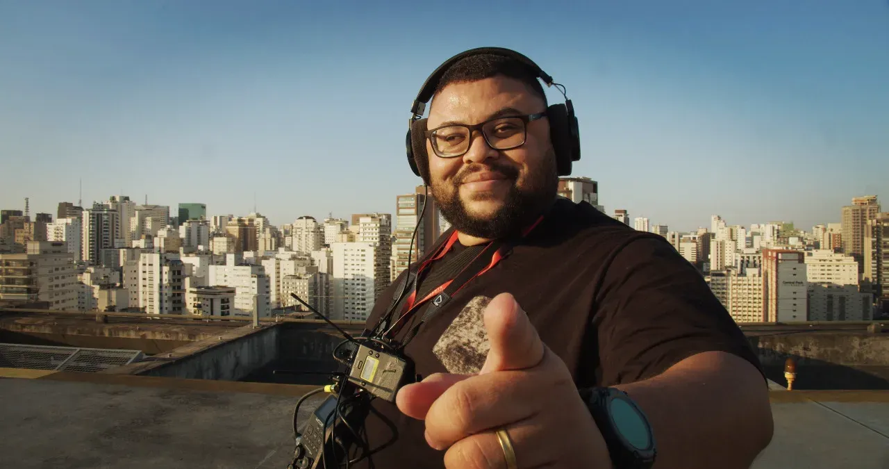 Maurilio, de fone e com o gravador de som direto no peito, aponta para a câmera em um terraço com os prédios de São Paulo ao fundo